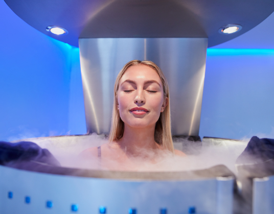 A blonde, white young woman appearing to enjoy standing in a futuristic metal tank up to her shoulders, with mist flowing around the opening